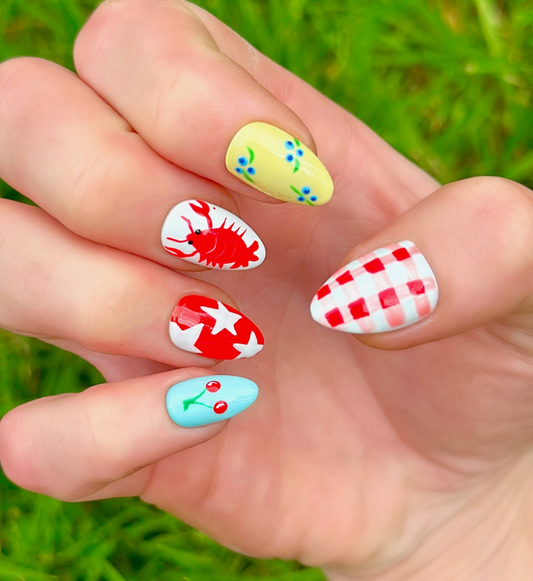 One hand displaying summer press on nails with vibrant, fun designs. Nail designs include a lobster, stars, cherries, gingham, and blueberries. Nail colors include red, white, orange, light blue, green, and yellow. The two hands are displaying the press on nails on a grassy summer background.