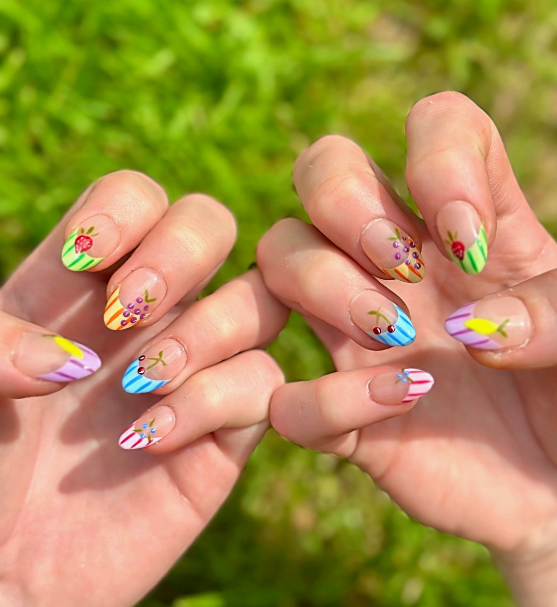 Two hands showing fruity Press On Nails with vibrant colors. Nails have colorful french striped tips and different fruits on each nail including grapes, cherries, lemon, blueberries, and strawberries. Hands are stacked on top of each other to display each press on design.