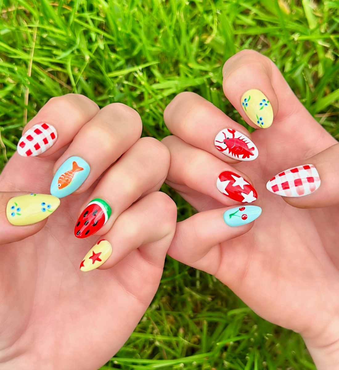 Two hands displaying summer press on nails with vibrant, fun designs. Nail designs include a lobster, stars, cherries, gingham, fish, watermelon, and blueberries. Nail colors include red, white, orange, light blue, green, and yellow. The two hands are displaying the press on nails on a grassy summer background.