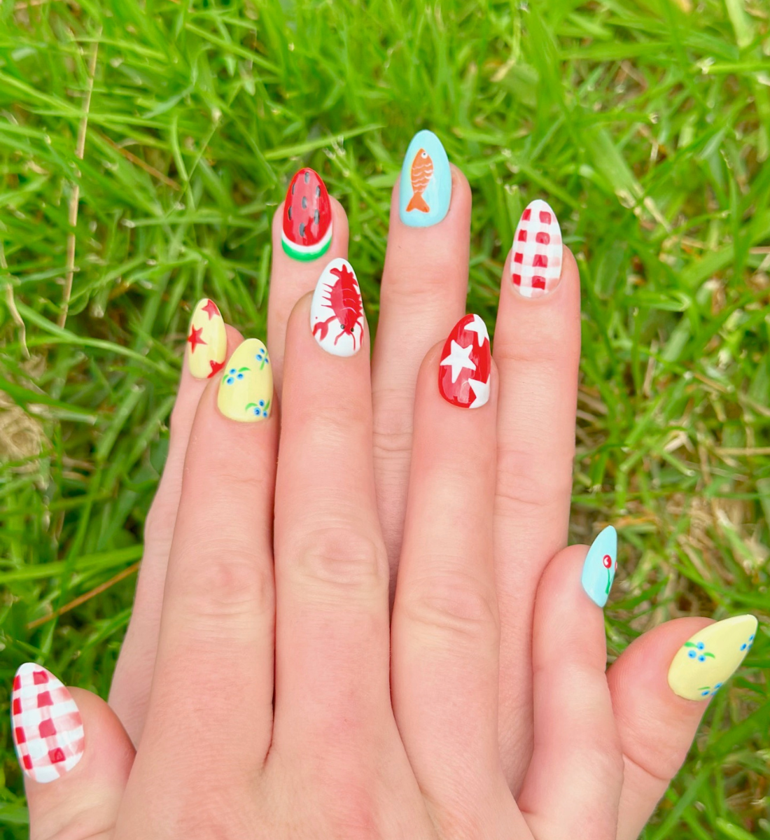 Two hands displaying summer press on nails with vibrant, fun designs. Nail designs include a lobster, stars, cherries, gingham, fish, watermelon, and blueberries. Nail colors include red, white, orange, light blue, green, and yellow. The two hands are displaying the press on nails on a grassy summer background.