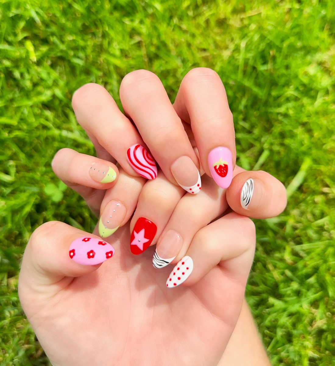 Hand-Painted acrylic press-on nails with vibrant colors and designs including strawberries, stars, gingham, zebra print, and french tips. The nail colors are red, light green, light pink, zebra print, and silver details. Hands and nails are displayed on grassy summer background.