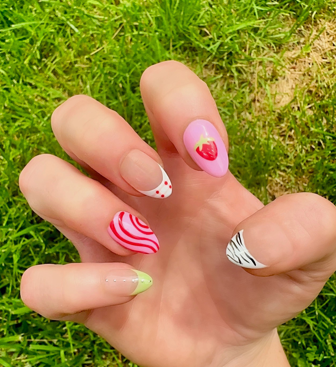 Hand-Painted acrylic press-on nails with vibrant colors and designs including strawberries, stars, gingham, zebra print, and french tips. The nail colors are red, light green, light pink, zebra print, and silver details. One hand displaying nails is on a grassy summer background.