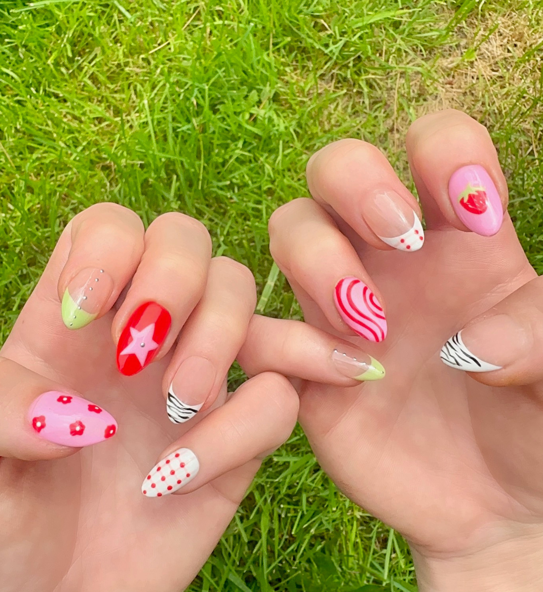 Hand-Painted acrylic press-on nails with vibrant colors and designs including strawberries, stars, gingham, zebra print, and french tips. The nail colors are red, light green, light pink, zebra print, and silver details. Two hands and nails are displayed on a grassy summer background.